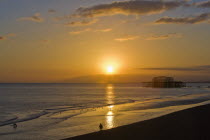 Dramatic orangy-red sunset silhouetting remains of the West Pier with dog and walker on the beach in the foreground