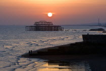 Dramatic orangy-red sunset silhouetting remains of the West Pier with beach and stone groyne in foreground