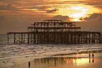 People walking along the wet sand at low tide at sunset  with a dramatic skyline  in front of the remains of the West Pier