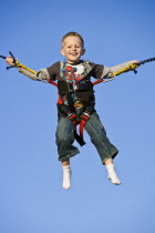 A young blond boy smiling while suspended in a harness on a seaside bouncy machine with a bright blue sky backround.
