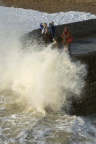 Waves crashing onto beach and groyne with people getting wet.