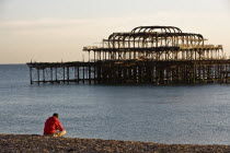 Single male with a hat and red windcheater sitting on the shingle beach with the rusty remains of the West Pier in the background.