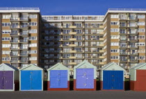 Multi-coloured beach huts on the tarmac promenade with a block of modern flats in the background.