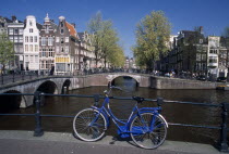 Blue bicycle leaning against a railing on bridge with Keizergracht Canal running parallel and Leidsegracht perpendicular. Traditional waterside house facades behind.