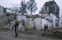 Woman carrying basket of vegetables on her head passing ruins of war damaged building.
