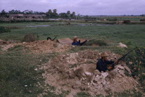 North Vietnamese combat troops  positioned with rifles and machine gun in dug-outs with paddy fields and workers behind.