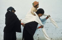 Indian woman helping another wrap young child in blanket sling on her back at Otavalo market.