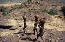 Dogon boys carrying water pots on their heads over rocky terrain.