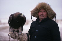 Kazakh man wearing thick  fur lined hat and holding eagle on his gloved hand.