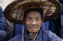 Head and shoulders portrait of elderly Bouyei man wearing wide brimmed woven hat.