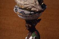 Woman on her way to market carrying laden bowls on her head and chewing on a teeth cleaning stick.