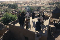 Members of ancient Dogon society performing rooftop dance holding carved staffs with view over surrounding village and landscape.