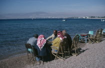 Group of men playing backgammon on shore of beach with Eliat and Israel in distance behind.