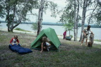 Canadian family on outback holiday setting up camp beside lake.  Children with tent and sleeping bag  mother with canoe and father using axe to split log.