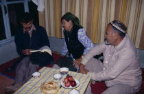 Uzbek family sat around tablke  with one man reading a book.