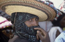 Portrait of Wodaabe man wearing wide brimmed hat and scarf over his head and mouth with hand raised to face.