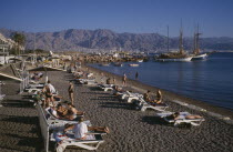Lines of sunbathers on white loungers on dark  sand beach with sailing ships moored against jetty behind and distant mountain range.