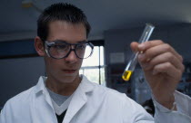 Male student wearing protective glasses and lab coat holding a test tube containing yellow liquid at an angle.