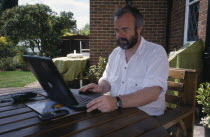 Man sitting at a table outdoors using a laptop linked to NFT via a mobile phone.