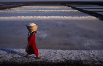 Woman wearing red carries salt in basket on her head across white salt pan