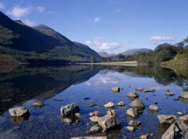 Buttermere.  Landscape  blue sky and drifting cloud reflected in lake with small rocks protruding from clear  shallow water in the foreground.