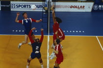 Indoor volleyball match with players at net leaping for ball.