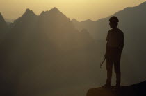 Climber in safety helmet standing in partial silhouette against mountain peaks in early morning light. France  Chamonix  Alps.