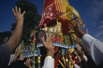 View over the reaching hands of a crowd toward brightly decorated cart with two people standing on the top during a festival