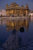 Sikh couple sitting on marble walkway looking out across sacred pool towards the Golden Temple and shimmering reflection on rippled water surface.Asia Asian Bharat Inde Indian Intiya Reflexion Religi...