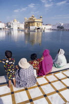 Women and children sitting on walkway beside sacred pool looking across to the Golden temple reflected in rippled surface of water.Asia Asian Bharat Female Woman Girl Lady Inde Indian Intiya Kids Rel...