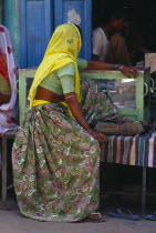 Woman in rose patterned sari and yellow head covering wearing ankle and toe ringsand bracelets  sitting outside jewellery shop. Asia Asian Bharat Female Women Girl Lady Inde Indian Intiya Jewelry One...