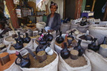 Female vendor behind display of sacks of cereals and pulses for sale at a market.