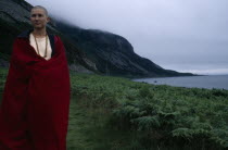 Tibetan Nun draped in a red robe standing next to coastline by green landscape and mountains at a Retreat on Holy Island.
