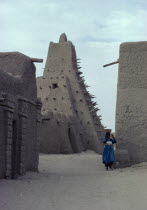 Street scene with mud built  Mosque and woman carrying a bowl on her head.
