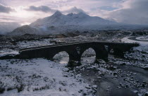 Sligachan and the Cuillin Mountains in winter snow.  Narrow bridge over frozen river and grey sky.