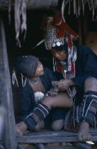Akha woman wearing traditional decorated silver head-dress and leggings sitting with child in hut entrance.