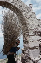 Taquile Island.  Local man carrying bundle of twigs on his back framed in partly seen stone archway.