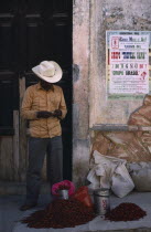 Man standing beside crumbling plaster wall with pile of red chillies for sale at his feet.
