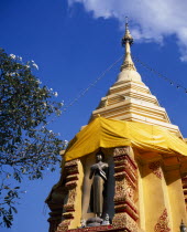 Standing Buddha statue below gold chedi wrapped in golden orange silk and hung with lights.Asian Prathet Thai Raja Anachakra Thai Religion Religious Siam Southeast Asia Siamese