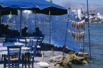 Restaurant outside seating with blue table and chairs next to waters edge with a fresh catch of seafood hanging from polesEuropean Ellada Greek Southern Europe