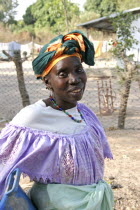 Tanji village.  Three-quarter portrait of woman wearing traditional head tie or scarf and bead necklace posing in front of her home compound. TanjehTanjihfaceAfricansmilecharacteristicsemotionexpressioncharacteristicsmoodcultureracejeweleryClassic Classical Female Women Girl Lady Gambian Historical Older One individual Solo Lone Solitary Western Africa