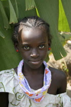 Tanji village.  Head and shoulders portrait of smiling young girl with braided hair and almond shaped eyes wearing patterned scarf tied around her neck and red earrings. TanjehTanjihAfricanfaceex...