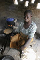 Tanji Village.  Smiling woman cooking traditional dish of groundnut sauce called mafay over open fire in kitchen which is outside of the house.  Sunlight shining through small  square openings in wall...