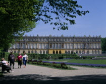 Chateau Herrenchiemsee. View across gardens to chateau with visitors walk on paths. Trees to the left and clear blue sky.