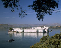 The Lake Palace framed by tree branches with white reflections on water and a boat travelling past