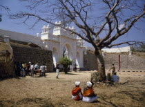 Maharaja s Palace.  Exterior wall and white  carved and decorated entrance arch with people gathered in dry  grassy area in foreground.