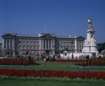 Buckingham Palace and the Victoria Memorial with crowds of visitors outside.  Formal flowerbeds with red and blue flowers in the foreground.