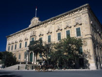 Auberge de Castille et Leon. Decorative baroque facade  with green doorway and shutters. Horse and carriage travelling past. Maltese flag flying from roof. Official residence of the Prime Minister