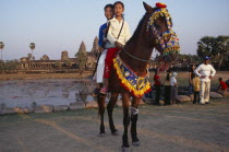 Cambodian children visiting Angkor during the Chinese New Year having photographs taken on pony wearing decorative harness in front of lake and temple complex.Pony has injury on near foreleg wrapped...