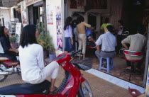 Busy hairderssers shop with two women sitting on mopeds watching a television in the back of the shopAsian Cambodian Kampuchea Southeast Asia Store 2 Female Woman Girl Lady Kamphuchea Female Women Gi...